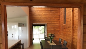 Un interior de madera con una mesa y sillas de comedor, una zona de cocina con armarios blancos, y vistas a través de un portillas de vidrio.