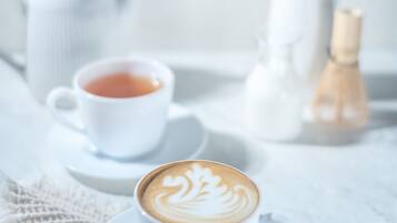 Una taza blanca de café con arte latte sobre un plato, una tetera blanca y una jarrón blanco sobre un mantel blanco.