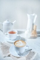 Una taza blanca de café con arte latte sobre un plato, una tetera blanca y una jarrón blanco sobre un mantel blanco.