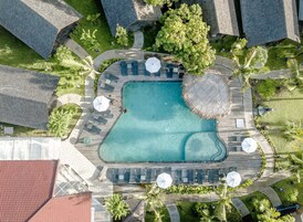 Vue aérienne d’un complexe hôtelier doté d’une piscine centrale, entourée d’espaces de détente avec des parasols, et agrémenté de diverses plantes tropicales.