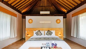 A hotel room with a wooden ceiling, a large bed with white bedding, two wall-mounted lamps, a bedside table with a tray of toiletries, and a wooden bench.