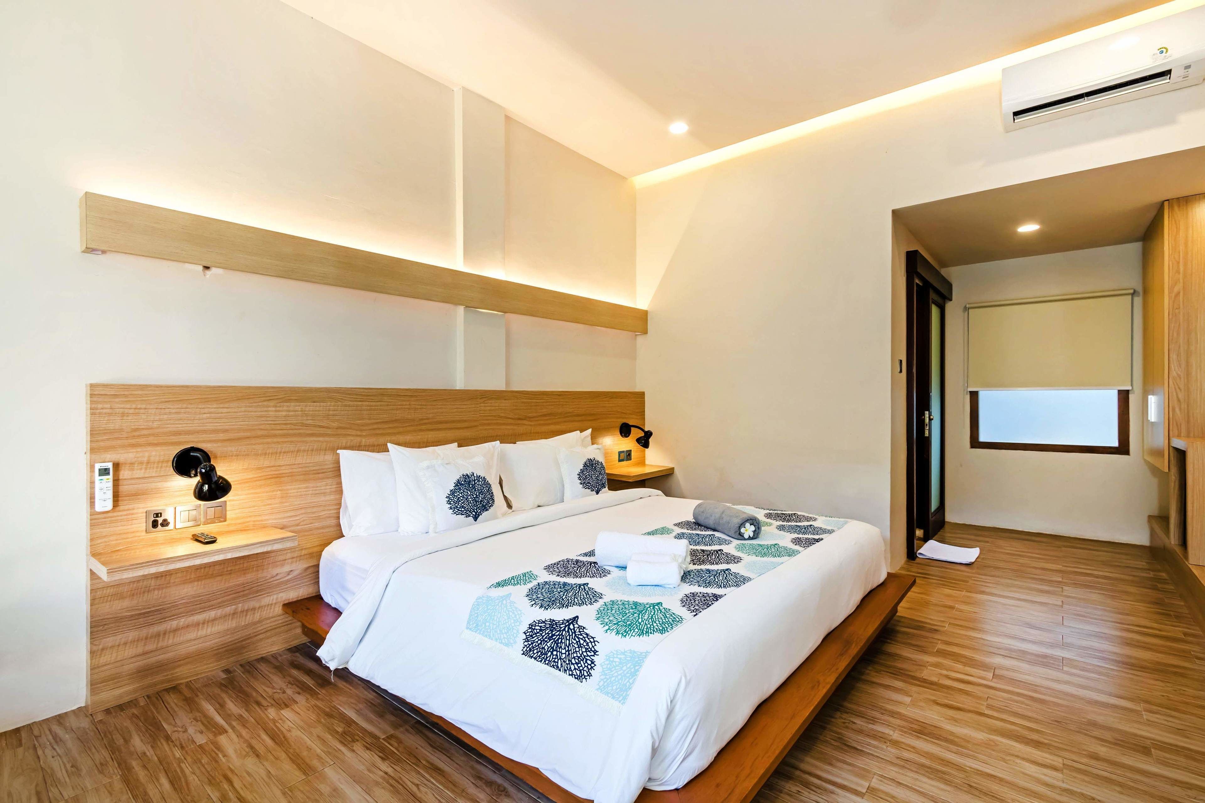 Une chambre d’hôtel moderne avec une tête de lit en bois, une table de chevet avec une lampe, un lit recouvert d’une couvre-lit à motifs géométriques et une grande fenêtre offrant une vue dégagée.