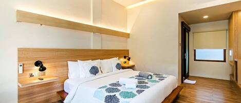 Une chambre d’hôtel moderne avec une tête de lit en bois, une table de chevet avec une lampe, un lit recouvert d’une couvre-lit à motifs géométriques et une grande fenêtre offrant une vue dégagée.