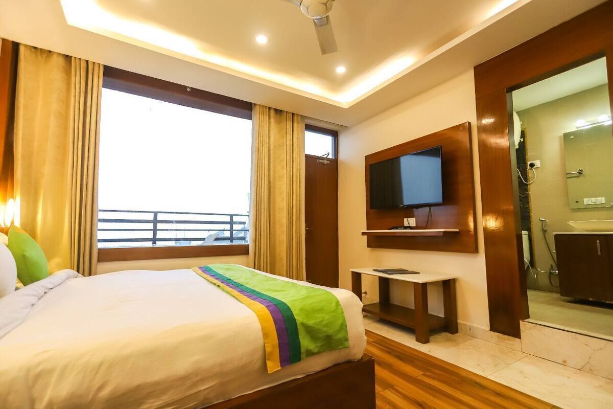 Quarto de hotel com cama, televisão, mesa pequena, banheiro com espelho e vista para a varanda.
