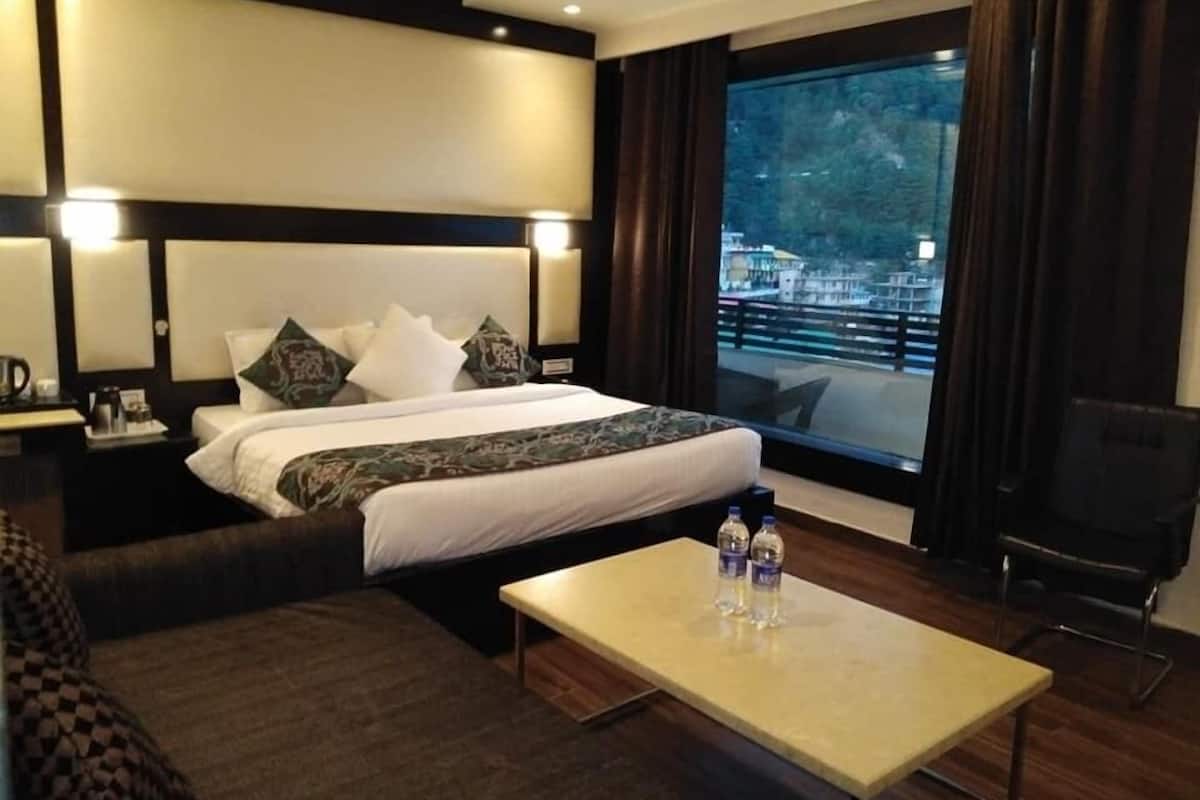 Quarto de hotel com uma cama grande, mesa de cabeceira com abajur, cafeteira, televisão e vista para a paisagem urbana através de uma grande janela.
