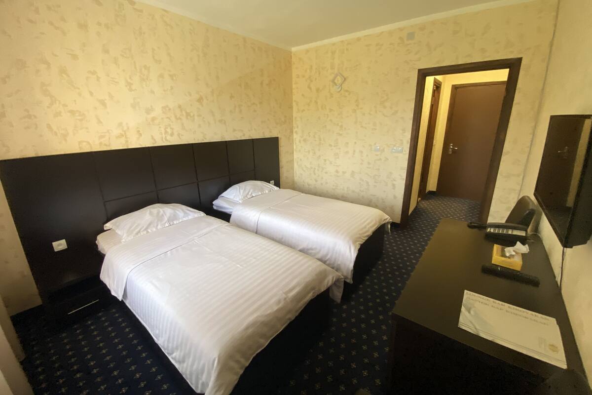 Quarto de hotel com duas camas, uma mesa de trabalho com telefone e uma porta que dá para outro quarto.