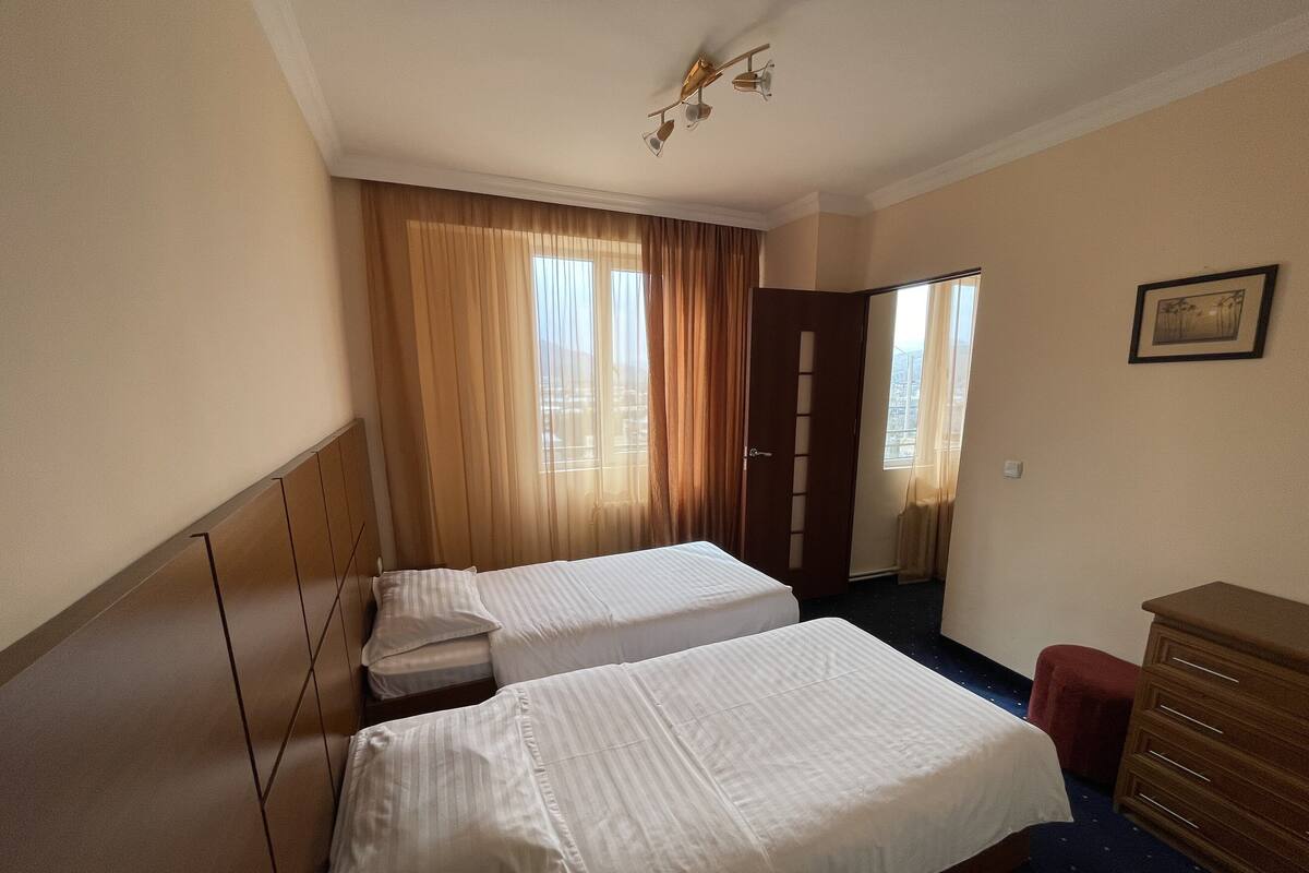 Quarto de hotel com duas camas, um aparador de madeira, uma cadeira vermelha e uma janela grande com cortinas.