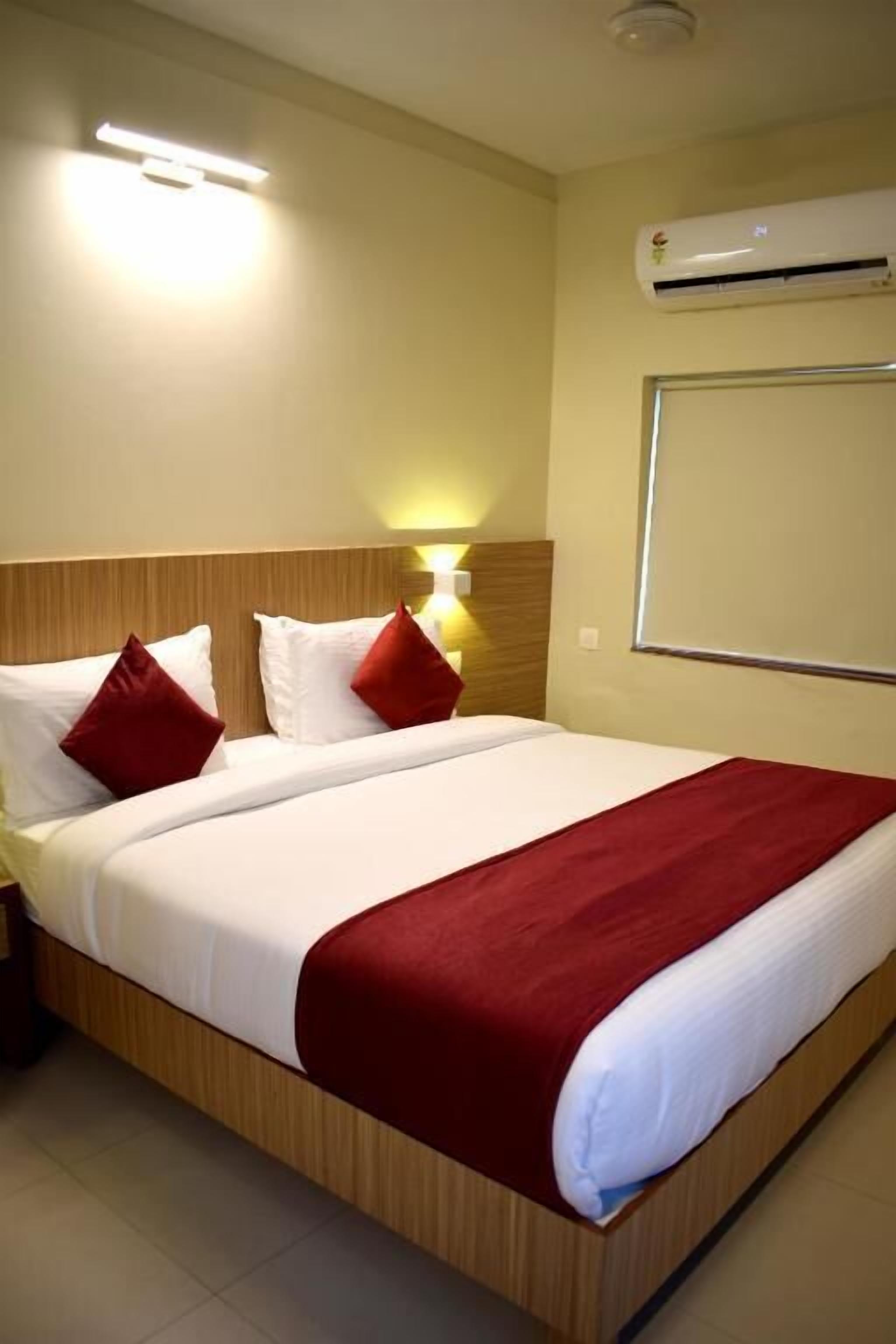 Ein Hotelzimmer mit hölzernem Kopfteil, einem großen Bett mit weißer und roter Bettwäsche, einer Wand-Klimaanlage und einer Wandleuchte.