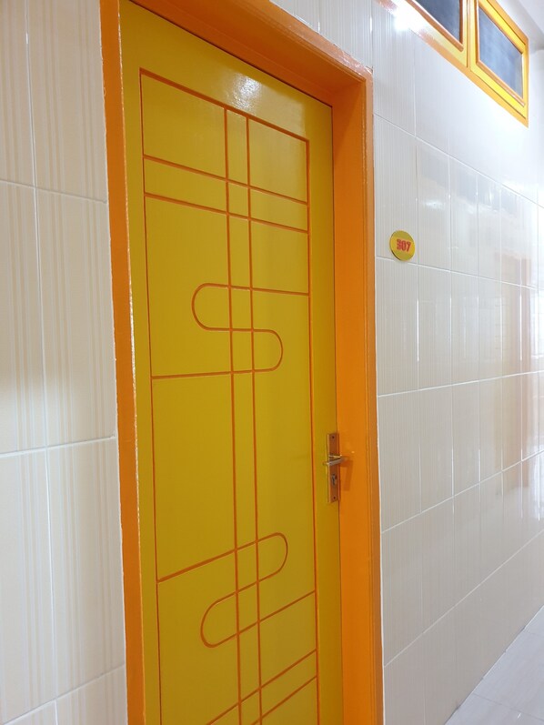Una puerta amarilla con diseño de cuadrícula y un pomo metálico, ubicada en un pasillo con paredes de azulejos blancos y una ventana con marco amarillo.