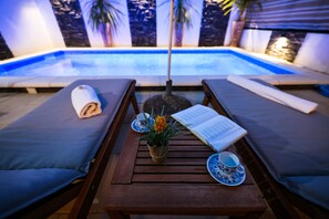 Un espace de détente aménagé au bord de la piscine, avec deux chaises longues, une table sur laquelle est posé un livre, un vase rempli de fleurs, ainsi qu’une tasse et une soucoupe.