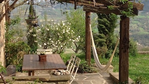 Un espacio de estar al aire libre rústico con una mesa de piedra y un banco, rodeado de vegetación y una hamaca.