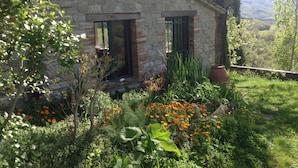 Un edificio de piedra con dos ventanas y una puerta, rodeado de exuberante vegetación y flores.