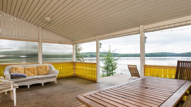 A covered outdoor deck with a wooden table and benches, a cushioned sofa, and a view of a lake. - Joutsen by Interhome (Kouvola)