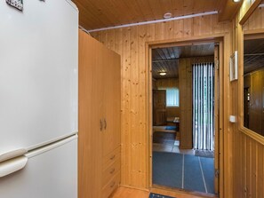 A wooden interior with a white refrigerator, a wooden wardrobe, and a door leading to a room with a bed and a window.