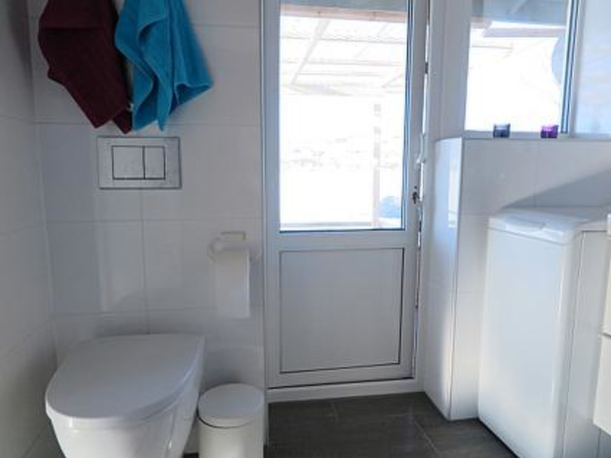 Un baño con inodoro, un cubo de basura y una puerta que da a una zona de lavandería.