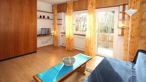 Una stanza con pavimento in legno, un divano blu, un tavolino con un cesto, una TV su uno scaffale e ampie finestre con tende.