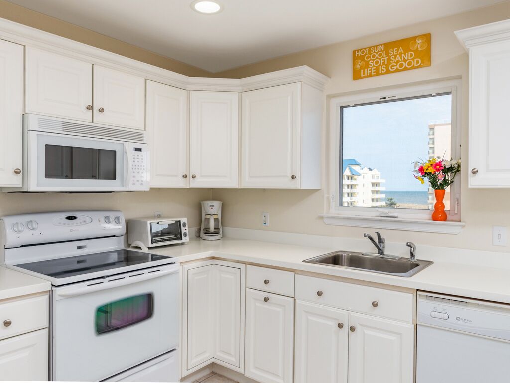 A kitchen with white cabinets, a microwave, a stove, a toaster, a sink, and a window with a view of buildings and flowers.