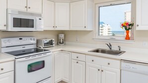 A kitchen with white cabinets, a microwave, a stove, a toaster, a sink, and a window with a view of buildings and flowers. - Beautiful 3 bedroom condo, mid town, ocean block (Ocean City)
