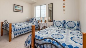 A bedroom with two single beds, a small table with lamps, a window with blinds, and a wall-mounted mirror. - Beautiful 3 bedroom condo, mid town, ocean block (Ocean City)