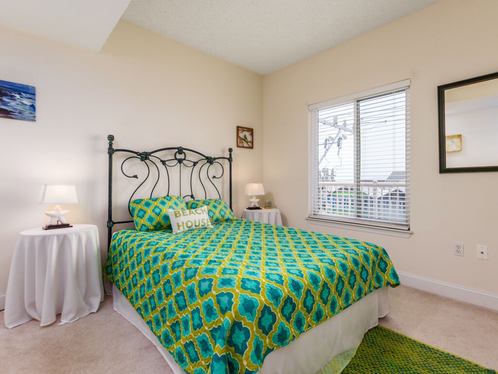 A bedroom with a bed, two bedside tables, a window with blinds, and a wall-mounted mirror.