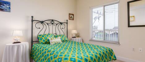 A bedroom with a bed, two bedside tables, a window with blinds, and a wall-mounted mirror.