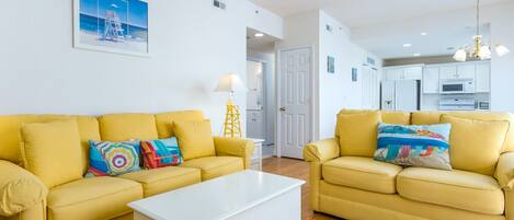 A living room with two yellow sofas, a white coffee table, and a kitchen in the background.