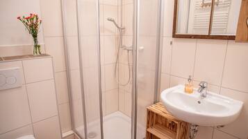 Un baño con ducha de vidrio, un lavabo blanco con dispensador de jabón y un taburete de madera.