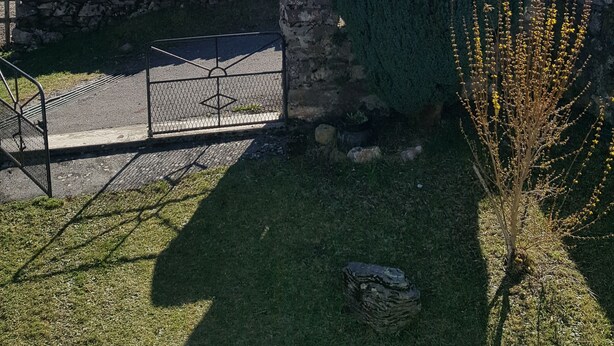 Un mur de pierres, un portail en métal et une zone herbeuse avec un arbre et un arbuste.