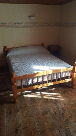 Un lit en bois avec une courtepointe dans une pièce dont le sol est recouvert de planches de bois et dont un des murs est décoré d’un motif.