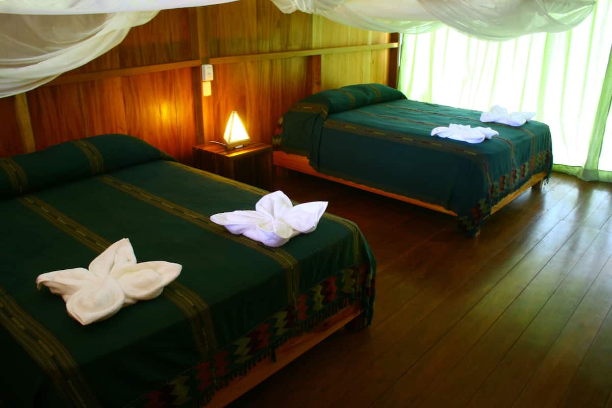 Two beds with green linens and white towels, a bedside lamp, and wooden walls.