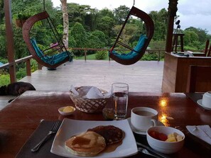 A dining area with a wooden deck, two hanging chairs, a table set with breakfast including pancakes, a glass of water, and a cup of coffee. - Finca Maresia (Drake Bay)