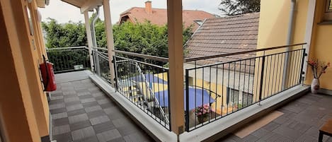 A covered balcony with a tiled floor, metal railing, and a view of a neighboring building with a red roof.