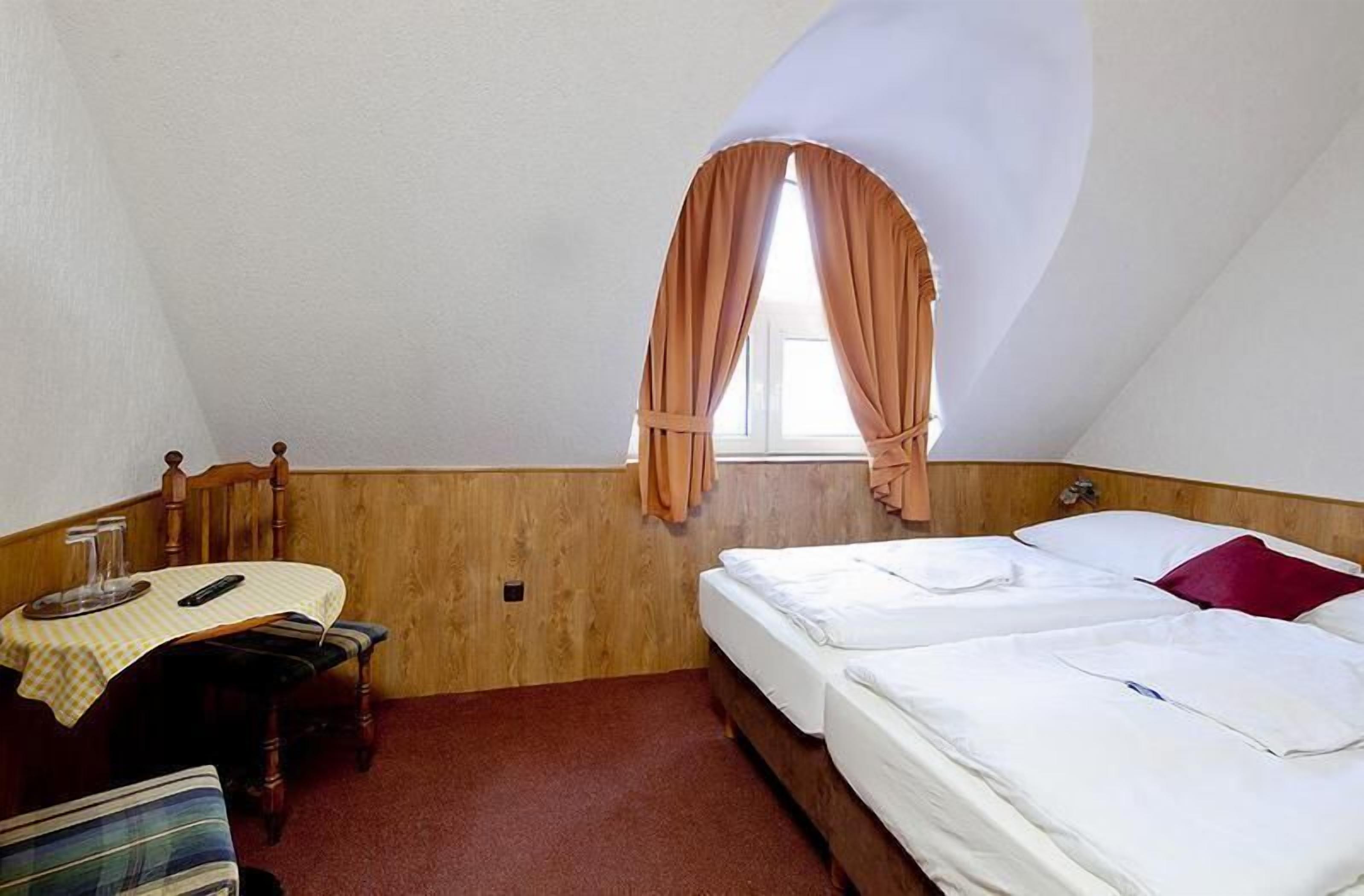 Una habitación pequeña y acogedora con dos camas, una mesa de madera con mantel a cuadros y una ventana con cortinas.