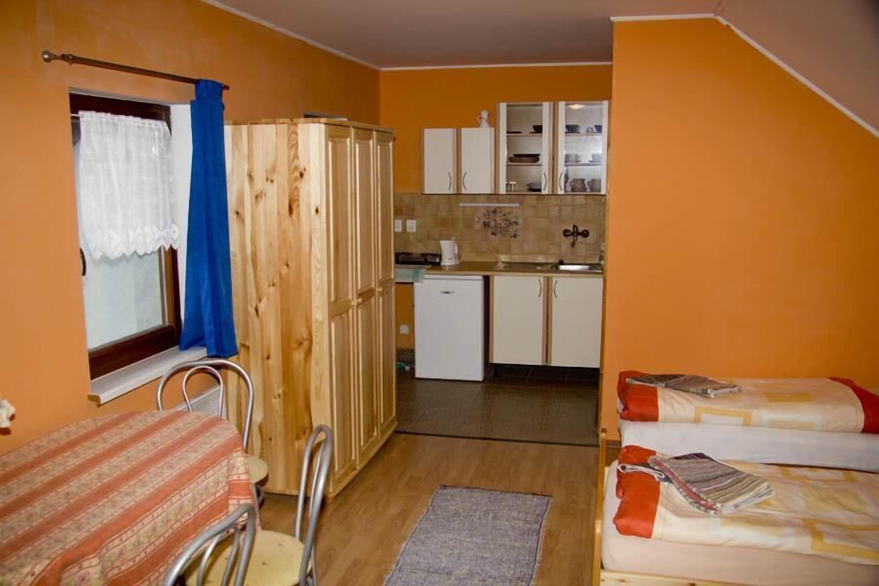 A compact room with a bed, a small table, a chair, a wooden wardrobe, a kitchenette with cabinets, and a window with curtains.