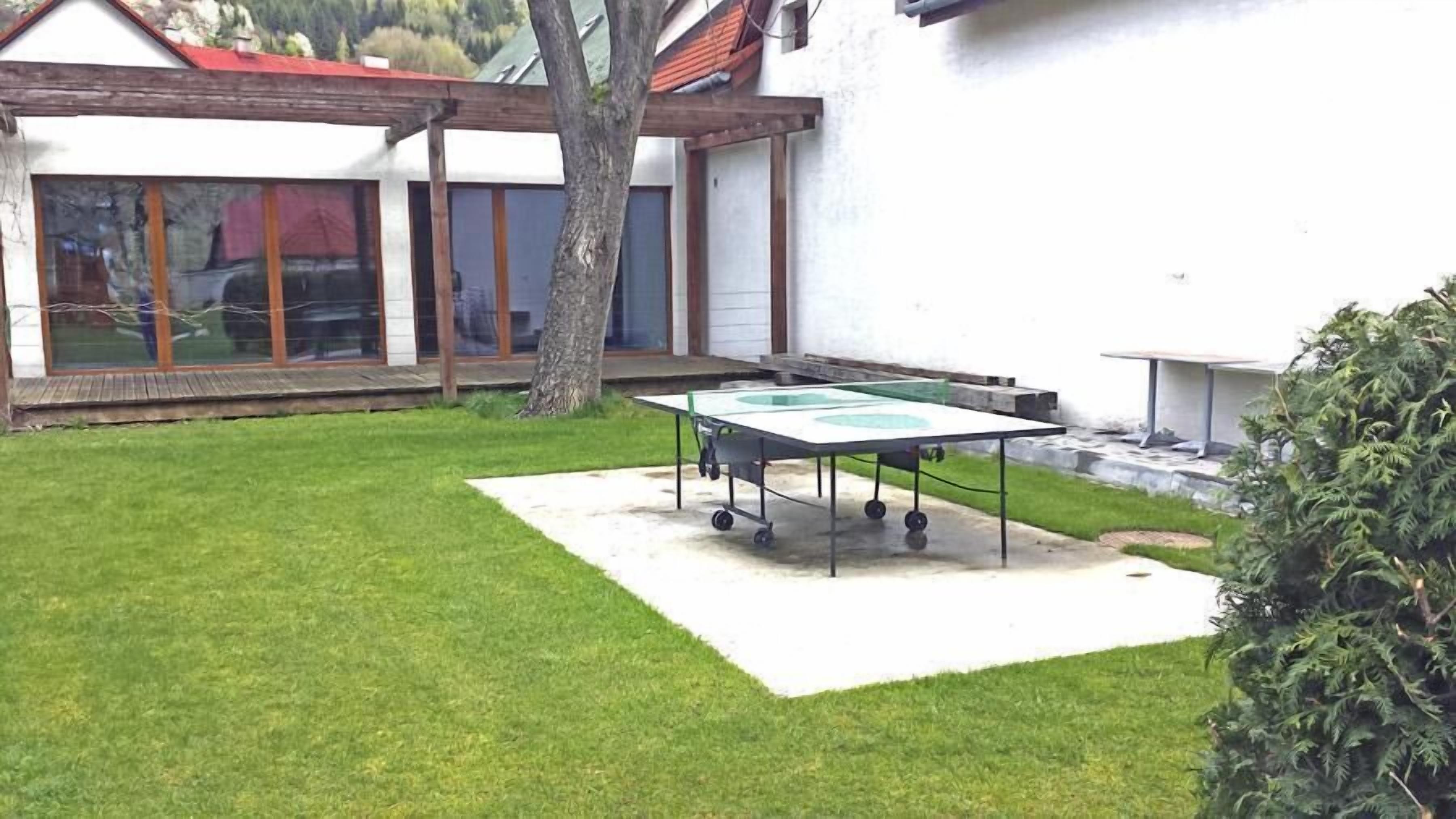 A small outdoor area with a ping pong table, a tree, and a building in the background.