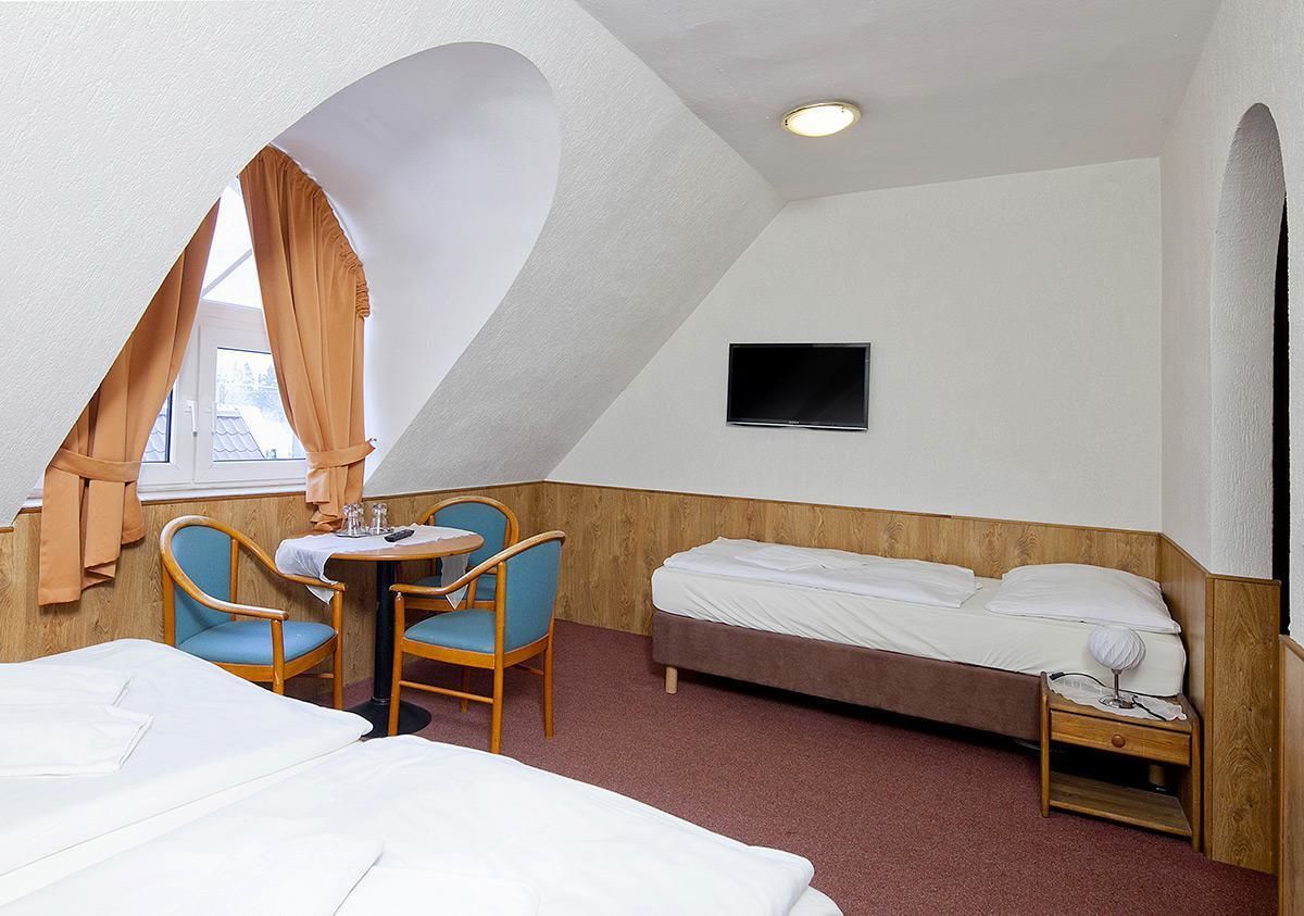 A hotel room with a bed, a desk with two chairs, a TV mounted on the wall, and a window with curtains.