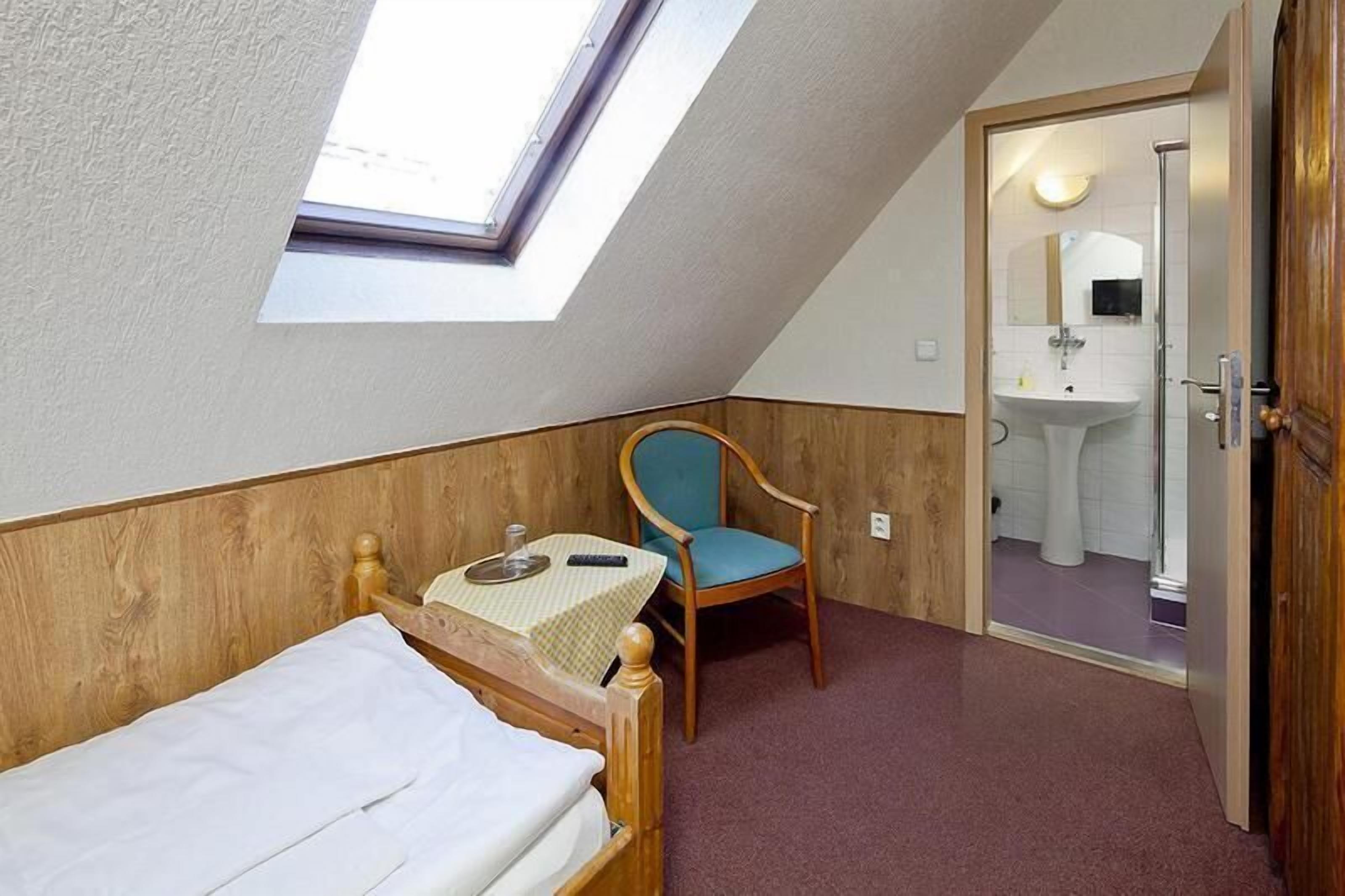 Una habitación pequeña y acogedora con techo inclinado, tragaluz, cama individual, una silla de madera y una mesita.