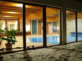A room with a large window overlooking a swimming pool, wooden ceiling beams, and a sandy floor.