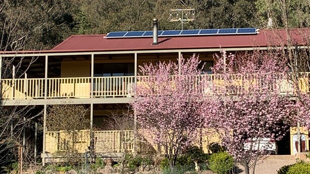 Ein zweistöckiges Gebäude mit rotem Dach und Solaranlage, umgeben von einem Garten mit blühenden Bäumen und Blick auf einen felsigen Berg.