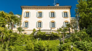 Un edificio grande e histórico con múltiples ventanas y un balcón, rodeado de abundante vegetación.