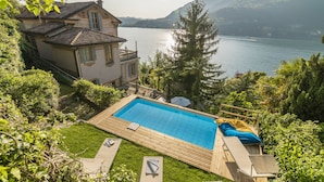 Una piscina con cubierta de madera, rodeada de exuberante vegetación y vistas a un lago y montañas.