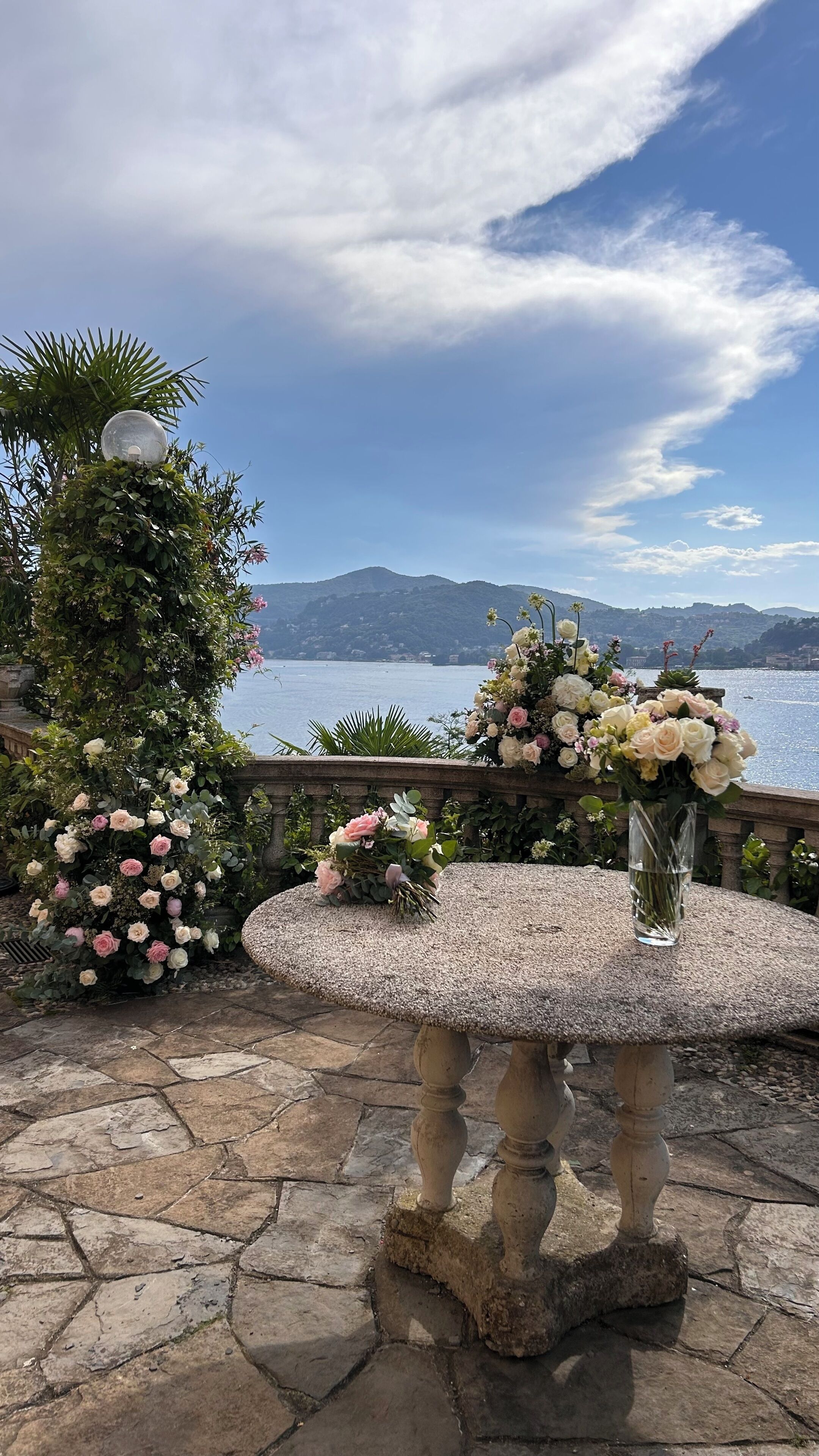 Ein Steintisch mit Blumen in einer Vase, ein dekorativer Bogen mit Kletterpflanzen und Blick auf einen See mit Bergen im Hintergrund.