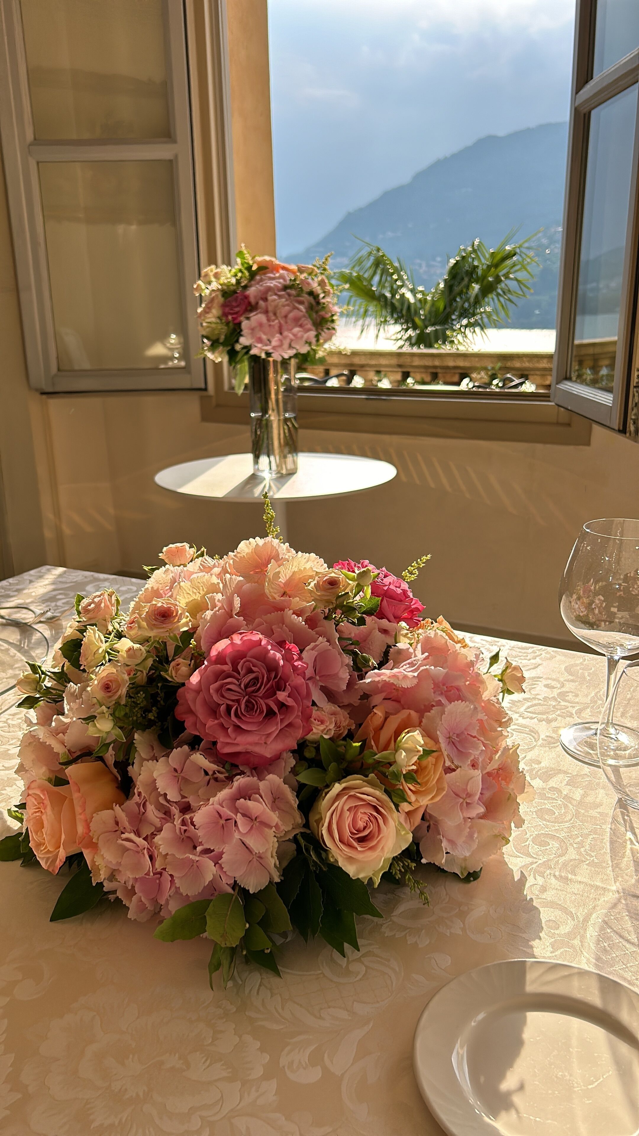 A table with a floral centerpiece, a glass of water, and a view of mountains through a window.