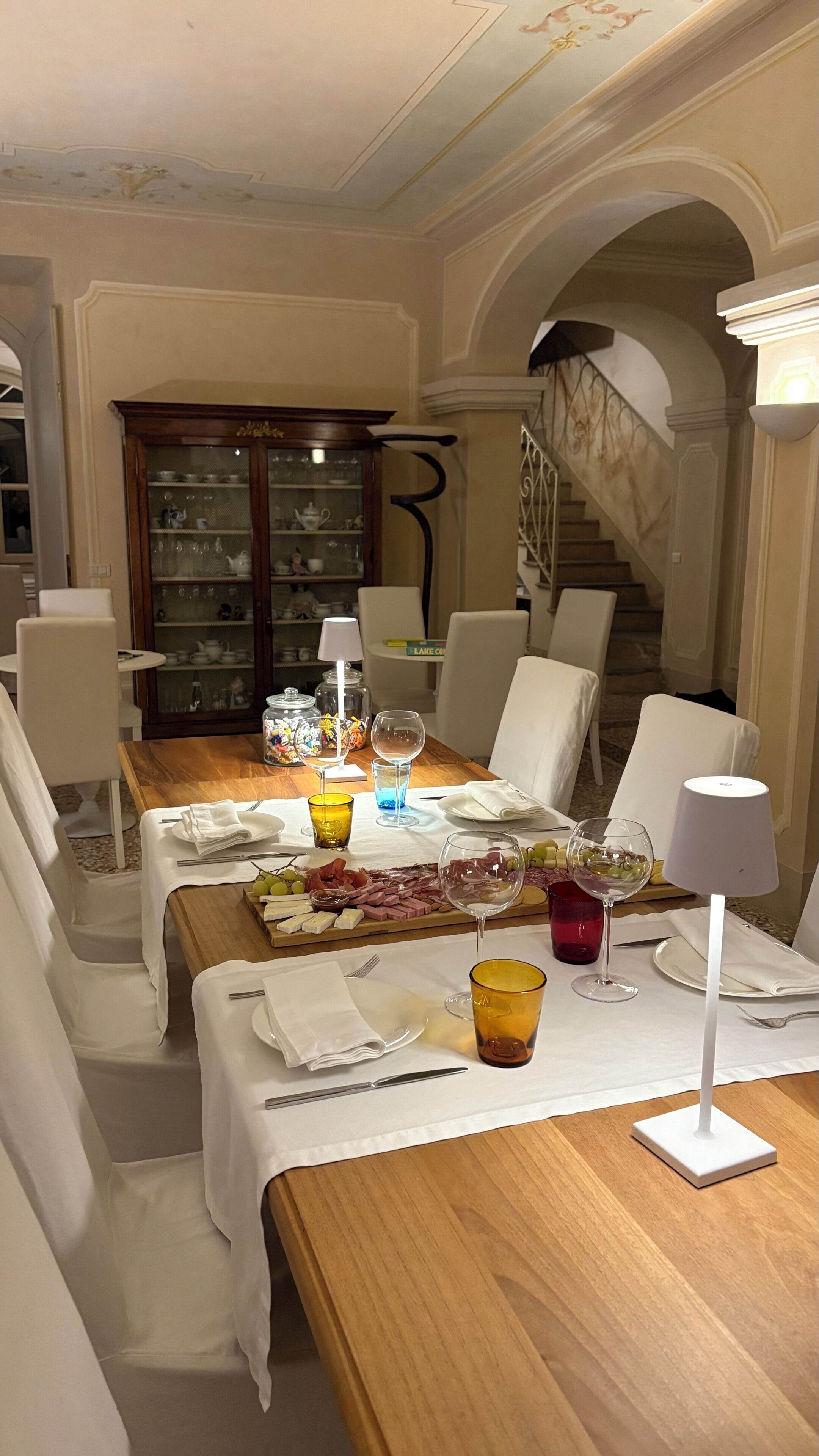 A dining area with a wooden table set for a meal, white chairs, a glass cabinet with dishes, and a staircase in the background.