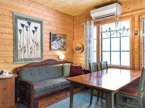 A wooden cabin interior with a sofa, dining table, chairs, a chandelier, and a painting.