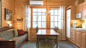 Ein gemütliches Holzhäuschen im Inneren mit Esstisch, Stühlen, einer Couch, einer Küchenzeile und einem großen Fenster mit Vorhängen.