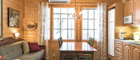 Interior de uma cabana de madeira com mesa de jantar, cadeiras, sofá, cozinha e uma janela grande.