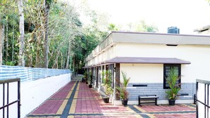 A corridor with a tiled floor, potted plants, a bench, and a building with a tiled exterior and a water tank on the roof. - City Holidays - Wayanad (Sulthan Bathery)