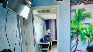 Una habitación con pared azul, un ventilador sobre una mesa, una cama con estructura blanca y un mural de palmeras.