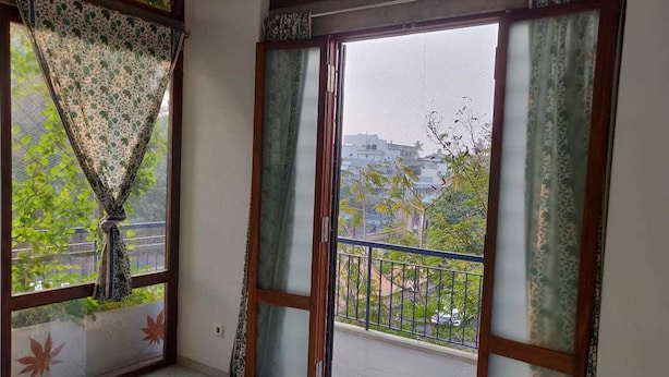 A room with a large window, a view of buildings and trees, and a balcony with a railing. - The Paperflower - A Wandertrails Stay (Puducherry)